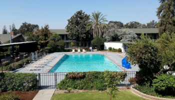 This image shows an outdoor swimming pool surrounded by a fence, lounge chairs, greenery, and a two-story building in the background.