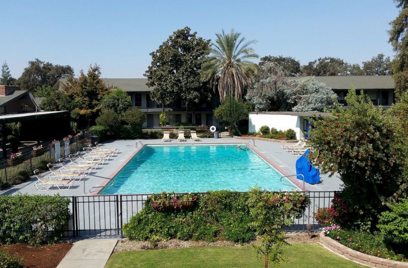 This image shows an outdoor swimming pool surrounded by a fence, lounge chairs, greenery, and a two-story building in the background.