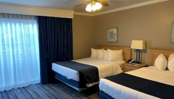 A hotel room with two beds, a nightstand with a lamp and phone, a ceiling fan, a large window with curtains, and framed artwork on the wall.