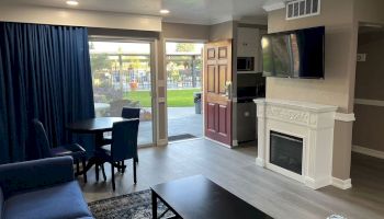 The image shows a living room with a sofa, coffee table, dining table, fireplace, TV, and an open door leading to an outdoor space with a lawn.