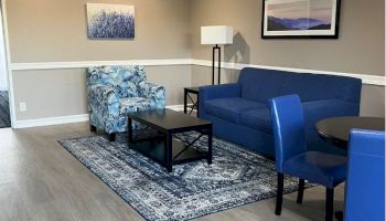 A cozy living room with a blue sofa, floral armchair, black coffee table, and matching blue chairs around a small dining table, decorated with art.