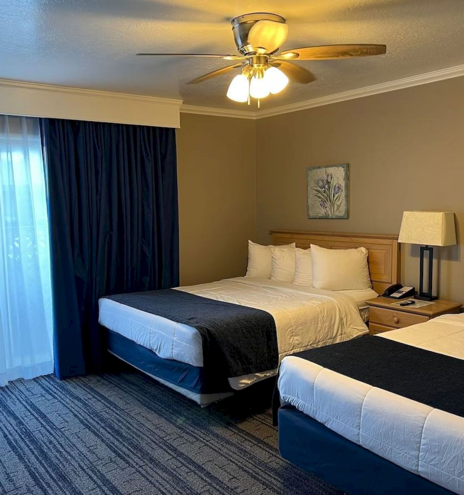 A hotel room features two double beds with dark and light beddings, a nightstand with a lamp and phone between them, and a ceiling fan above.