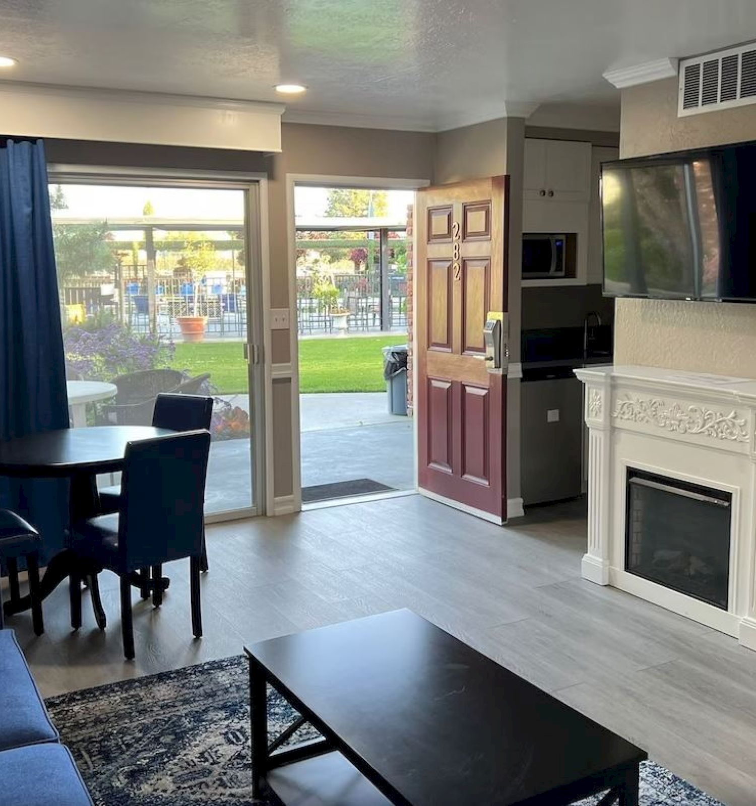 A living room with a fireplace, flat-screen TV, blue sofa, round dining table with blue chairs, and an open door leading outside, revealing a patio.