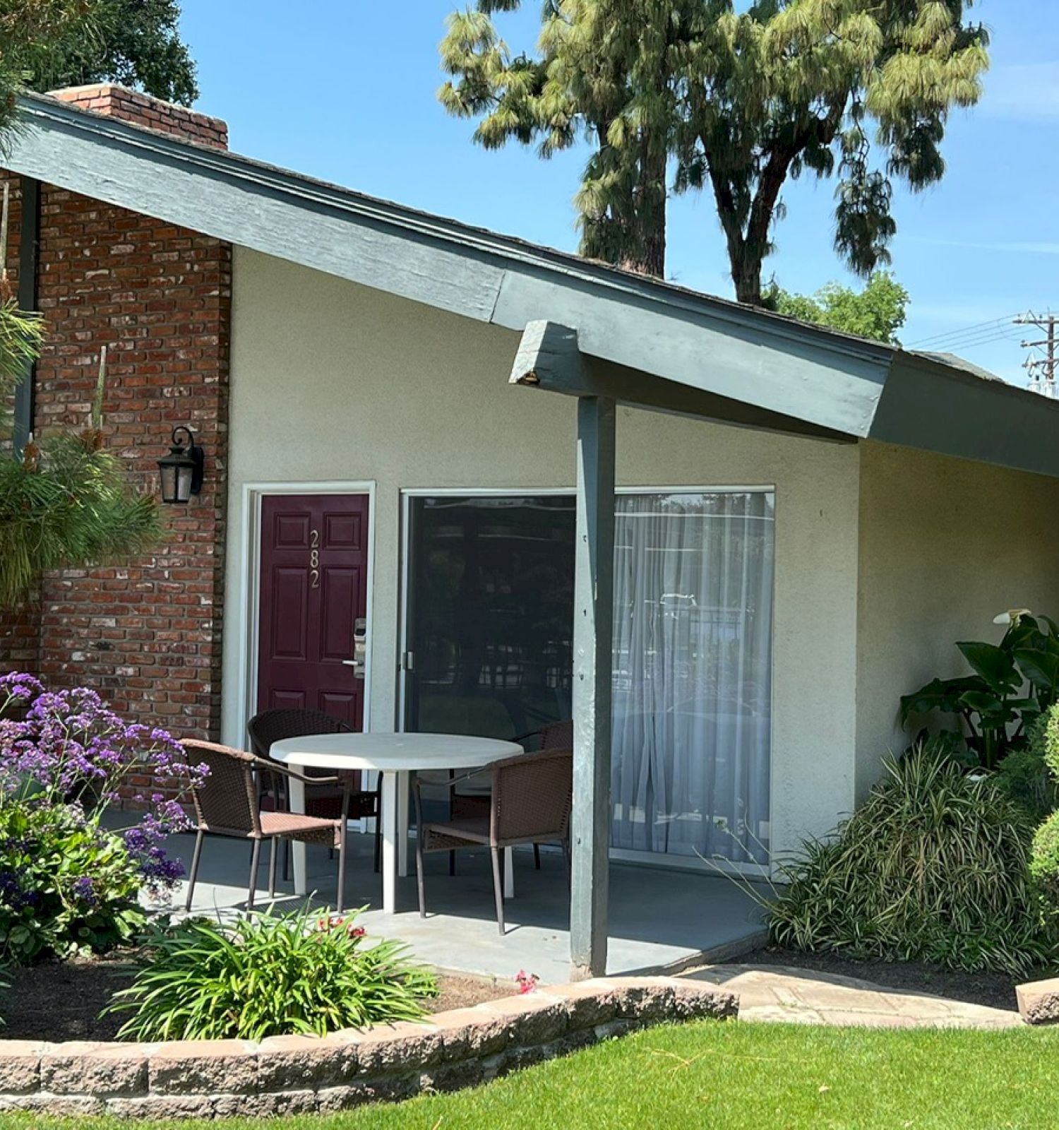 A small patio with table and chairs is attached to a house with a red brick wall, surrounded by green shrubs, flowering plants, and a well-maintained lawn.