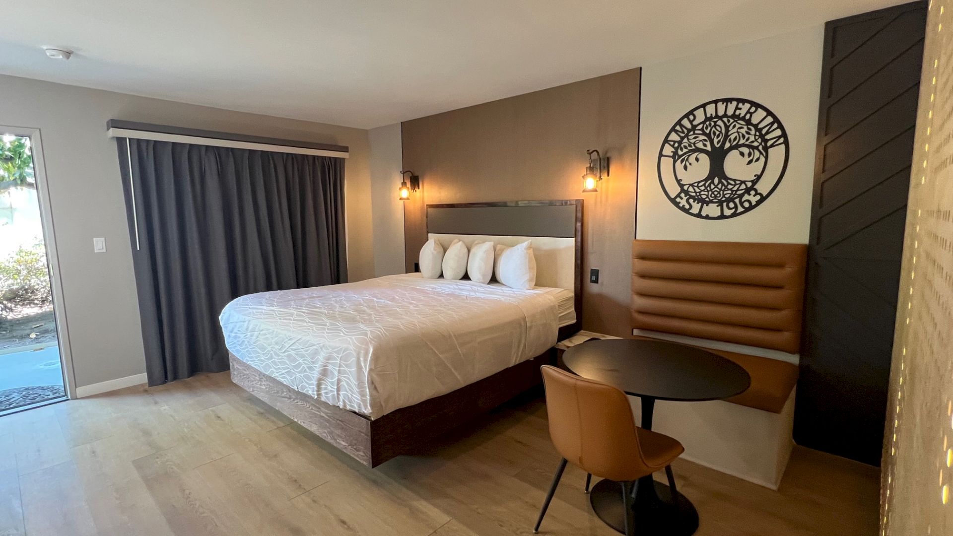 The image shows a modern hotel room with a bed, wall art, round table, chair, bench seating, and large curtained window.