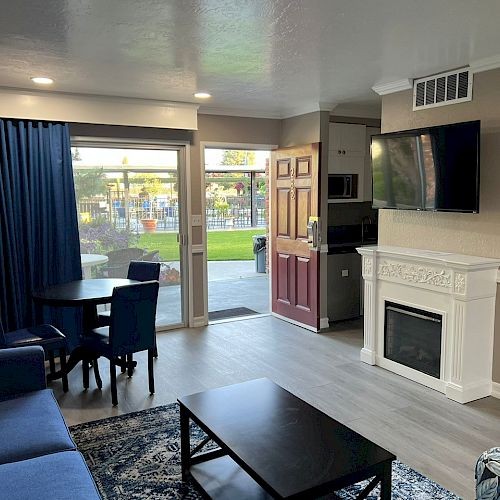 A modern living room with a fireplace, TV, blue sofa, coffee table, dining table, and large window overlooking a patio and garden.