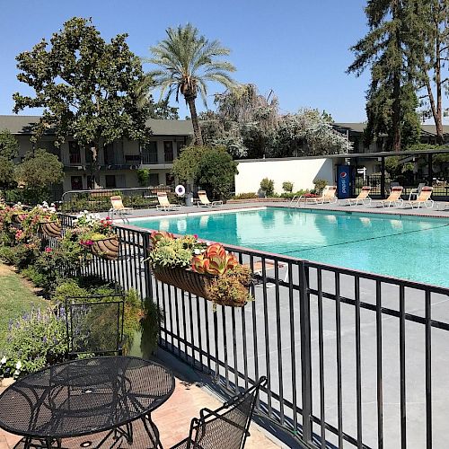 An outdoor pool area with lounge chairs, a fence adorned with flowers, a small seating area with tables, and trees in the background, on a sunny day.