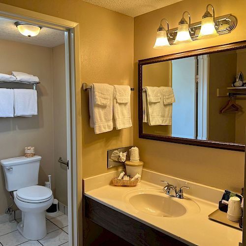 A well-lit bathroom with a large mirror, sink, and neatly folded towels. Adjacent room has a toilet, towel rack, and additional towels above.