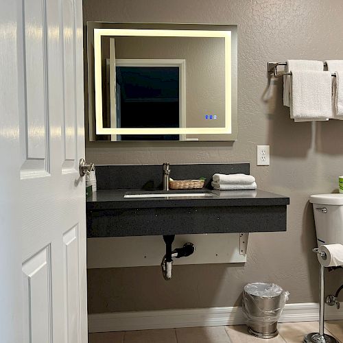 This image shows a bathroom with a modern design, featuring a backlit mirror, a black sink counter, a white toilet, and neatly folded towels.