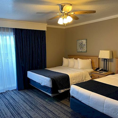 A hotel room with two beds, a ceiling fan, bedside table with phone and lamp, carpeted floor, large window with curtains, and wall art.