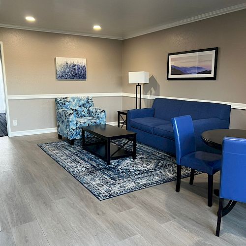 A cozy living room with blue furniture, a patterned rug, wall art, a floor lamp, and a round dining table with blue chairs on wooden flooring.