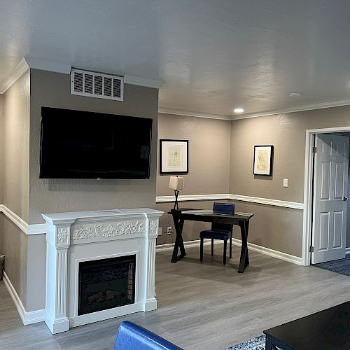 A modern living room with a wall-mounted TV, a fireplace, a desk with a chair, and framed pictures, featuring neutral colors and recessed lighting.