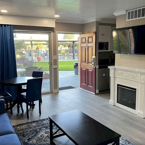 The image shows a cozy, modern living room with a sofa, coffee table, dining table, TV above a fireplace, and large windows with blue curtains.