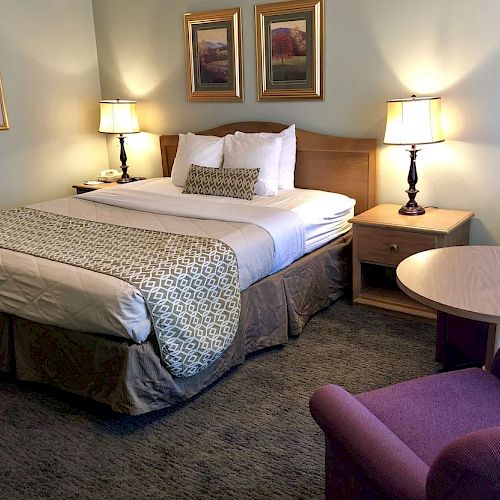 A neatly arranged hotel room with a double bed, two bedside tables with lamps, a small table with two chairs, and framed artwork on the walls.