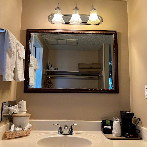 A bathroom sink with a mirror above, lit by three bulbs; towels, a coffee maker, cups, and toiletries are placed on the counter.