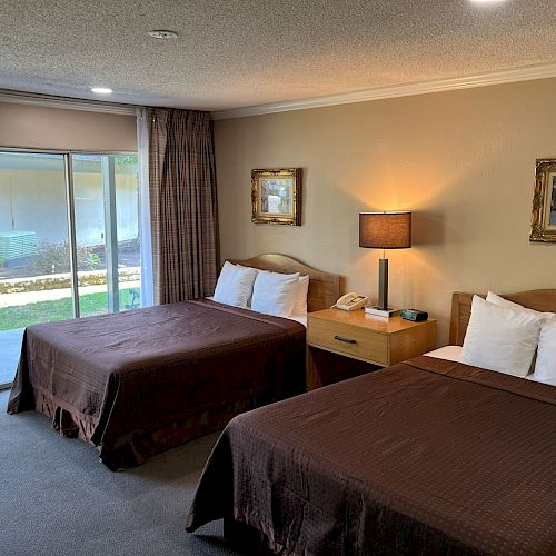 This image shows a hotel room with two beds, a bedside table with a lamp, two paintings on the wall, and a sliding glass door leading outside.