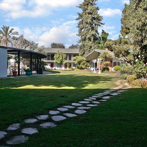 A green lawn with a stone path leads to a building surrounded by trees and plants under a blue sky with scattered clouds.