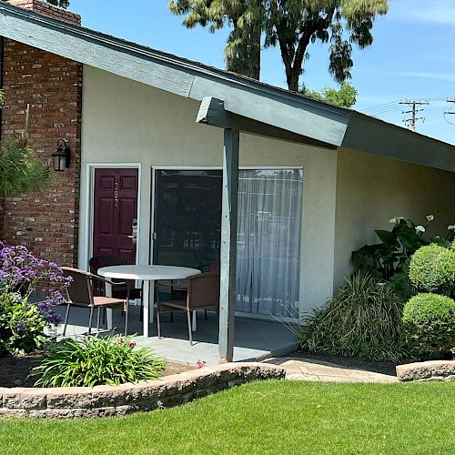 The image shows a house exterior with a patio, table, and chairs, next to a garden with neatly trimmed shrubs, flowers, and lush green grass.