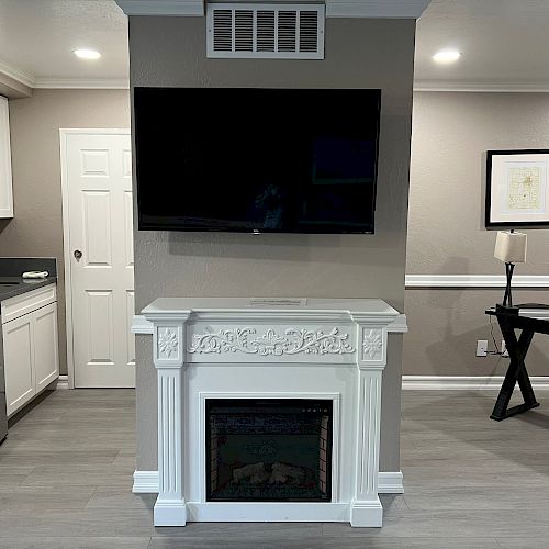 A modern living space with a mounted TV, fireplace, kitchen area, and a desk with a lamp and framed picture hanging above.