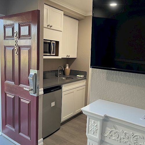 The image shows an open door to a room numbered 282, revealing a kitchenette with a sink, dishwasher, microwave, and cabinets, adjacent to a large TV.