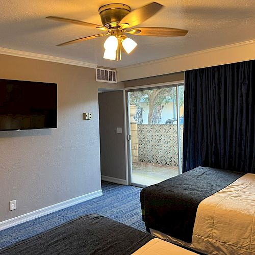 A hotel room with a ceiling fan, wall-mounted TV, two beds, blue curtains, and a sliding door leading to a patio or balcony ending the sentence.