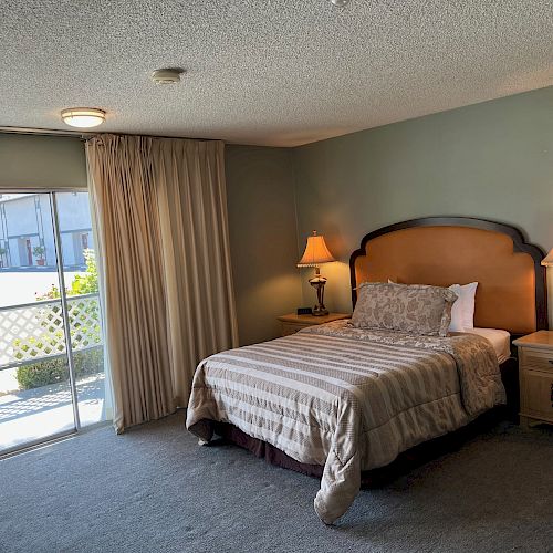 A neatly arranged bedroom with a queen-sized bed, bedside tables with lamps, a sliding door leading outside, and a large window with curtains.