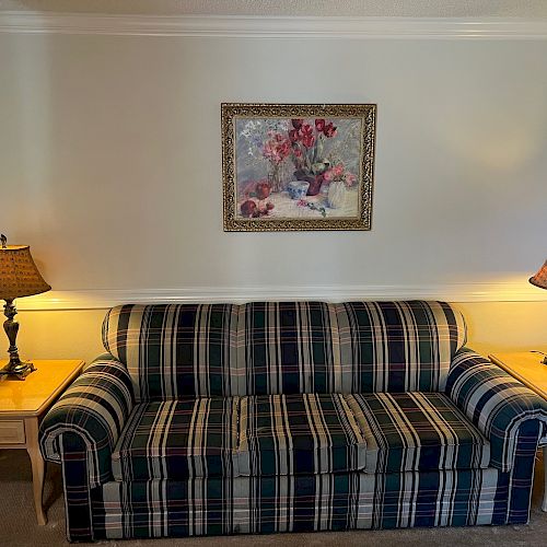 A living room features a plaid sofa, two side tables with lamps on either side, and a floral painting centered above the sofa.