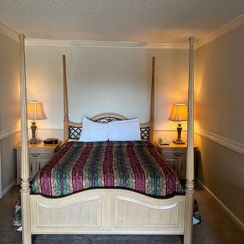 The image shows a neatly made bed with a colorful quilt, flanked by two nightstands, each with a lit lamp. A carpet covers the floor.