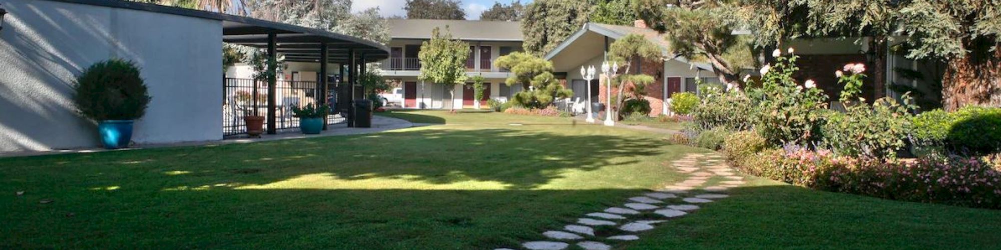 The image shows a lush garden with a stone pathway, surrounded by greenery, leading to a modern building with outdoor seating areas.