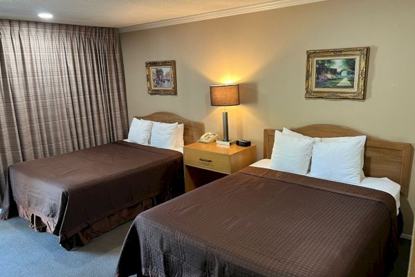 The image shows a hotel room with two double beds, a nightstand with a lamp and phone, framed artwork on the walls, and brown bedspreads and curtains.