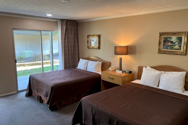 The image shows a hotel room with two double beds, brown bedspreads, a bedside table with a lamp, two paintings on the wall, and a sliding glass door.