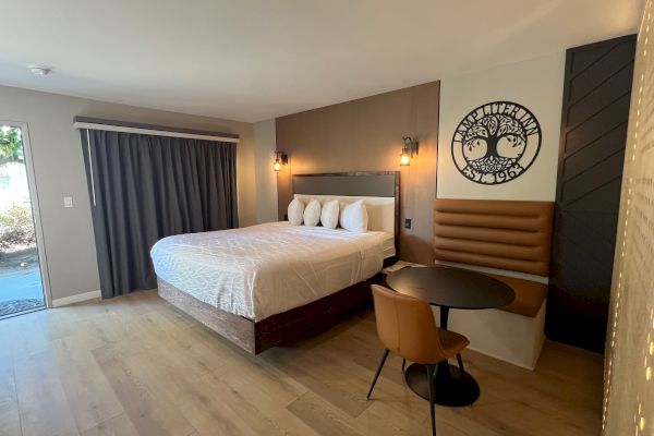 A modern hotel room features a bed, round table, and seating area, with wall art and curtains.
