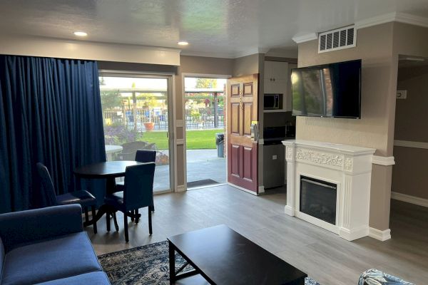 A modern living area with a sofa, dining table, fireplace, wall-mounted TV, and a glass door leading to an outdoor area.