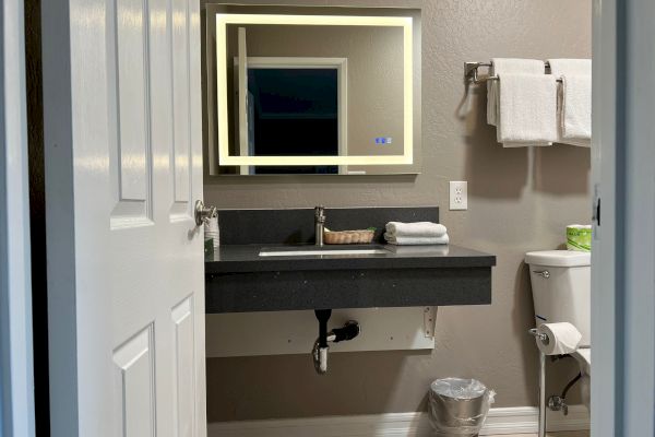 The image shows a bathroom with a wall-mounted sink, an illuminated mirror, a toilet, towels on a rack, a trash bin, and some toiletries.