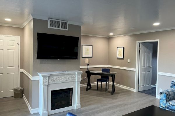 A modern living room with a wall-mounted TV, fireplace, desk, and chair, featuring art on beige walls and wood flooring, adjacent to a hallway.