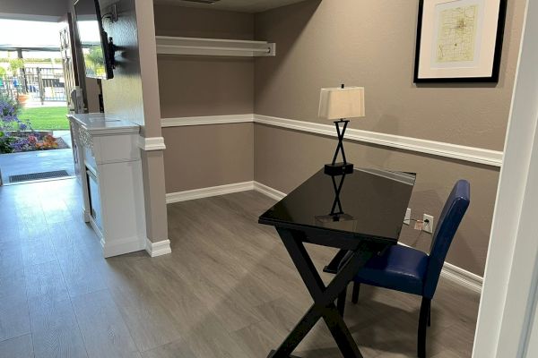 A small room with a black glass desk, blue chair, table lamp, wall art, white shelves, fireplace, and view of an outdoor area through the open door.