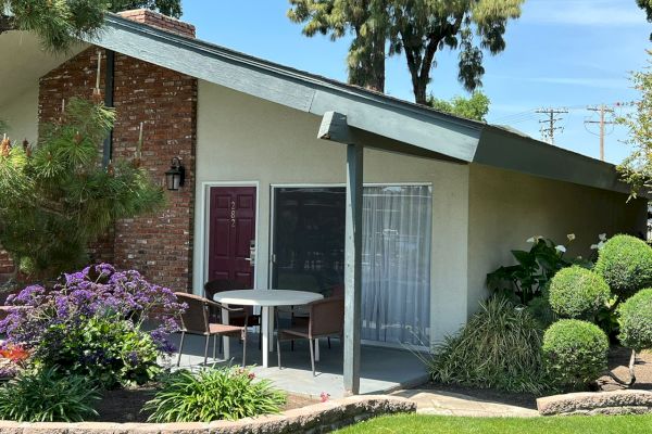 A small house with a front porch, table, chairs, and well-maintained garden featuring trimmed bushes, flowering plants, and a lush lawn.