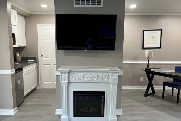 A modern living room with a flat-screen TV above a decorative fireplace, a kitchen on the left, and a desk with a lamp and chair on the right.