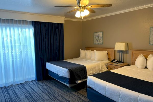 The image shows a hotel room with two double beds, each with white bedding and dark blue runners, a ceiling fan, a lamp, and floor-to-ceiling windows.