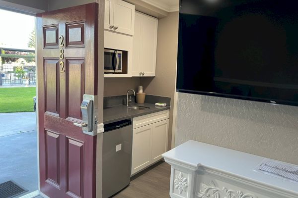 The image shows an open door numbered 292 leading to a room with a kitchenette, including a microwave, sink, dishwasher, TV, and drawers.