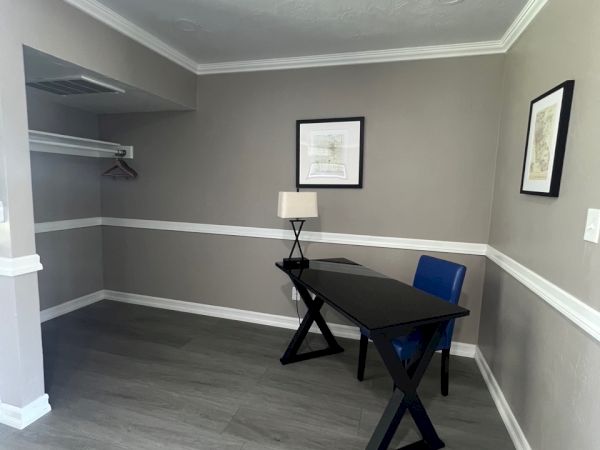 A small, minimalist room with a black desk, blue chair, lamp, and two framed pictures on gray walls. There's a closet with a hanger.