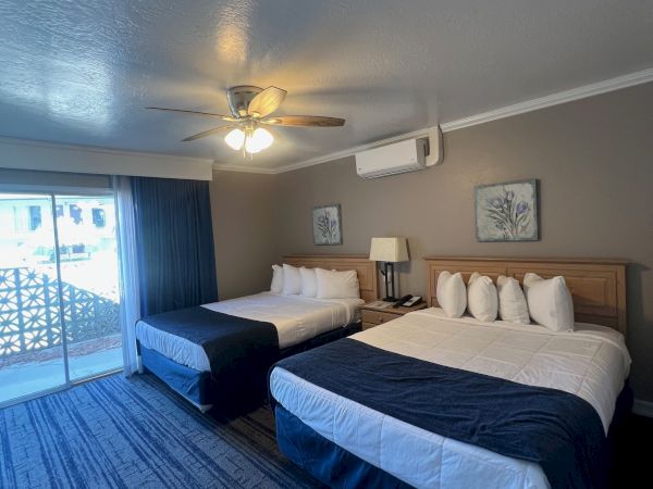 A hotel room with two large beds, decorative pillows, bedside lamps, and a sliding door to a balcony, under soft lighting.