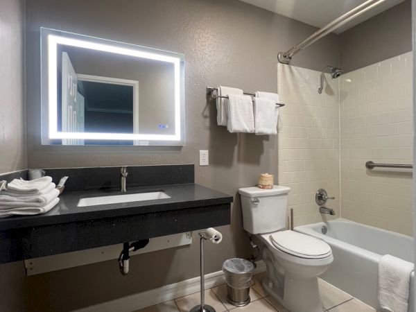 A modern bathroom with a lit mirror, sink with towels, toilet, and bathtub with a shower curtain in a neutral color scheme.