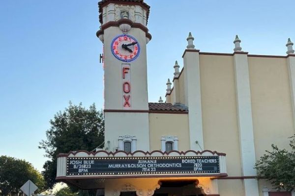 An exterior view of the Fox Theater, showing its iconic tower, clock, and marquee. Several people are gathered outside the entrance.