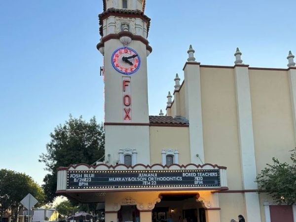 An exterior view of the Fox Theater, showing its iconic tower, clock, and marquee. Several people are gathered outside the entrance.