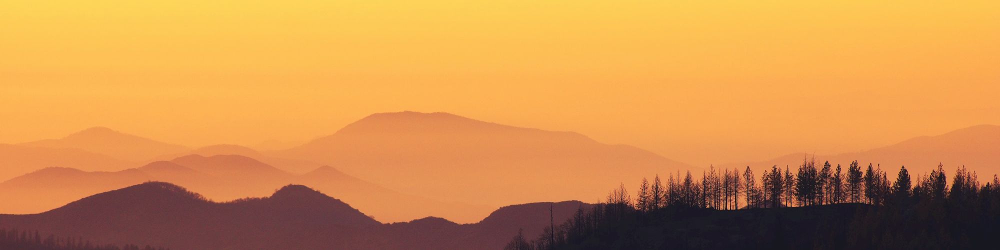 A beautiful landscape image featuring mountains against a vibrant yellow-orange sky at sunset, with trees silhouetted in the foreground.