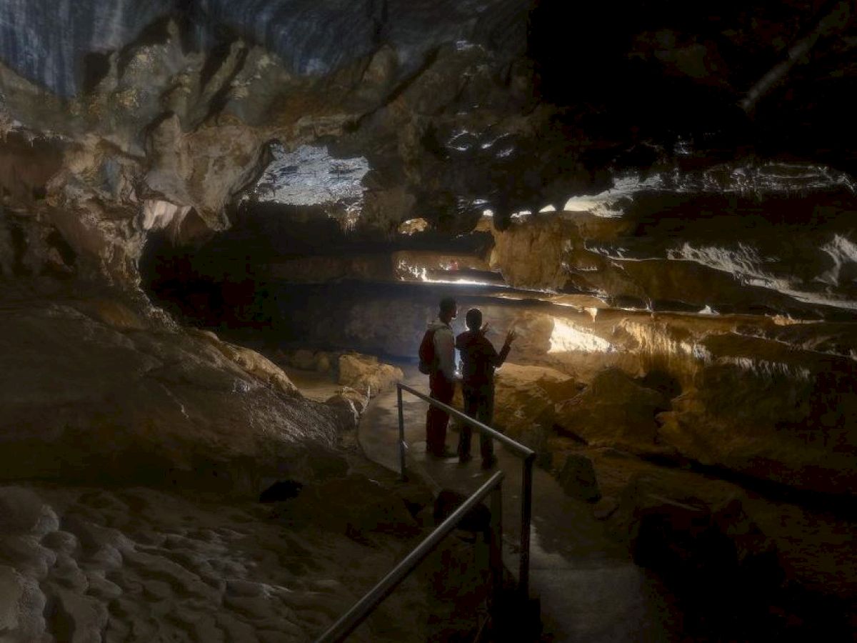 Two people explore a dimly lit cave with stalactites and stalagmites. They appear to be standing on a pathway railing.