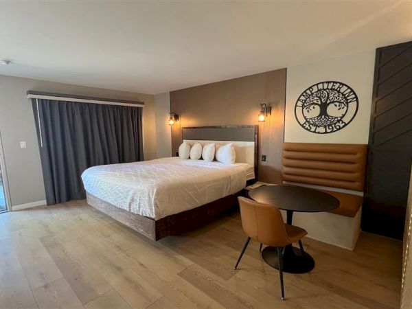 A modern hotel room with a large bed, side table, chair, and wall art, featuring wooden flooring and large drapes by the window.