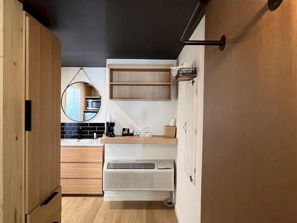 The image shows a modern interior with a wooden floor, shelving, closet space, and a small kitchenette area with a mirror and sink.