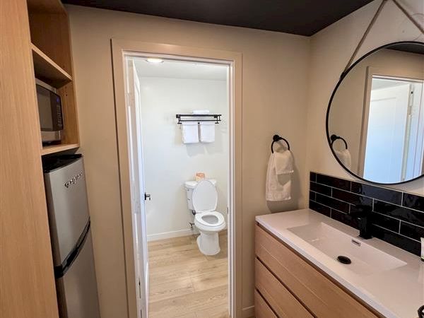 A small bathroom features a wooden vanity, round mirror, and visible toilet through a doorway, with light wood floors and a dark ceiling.